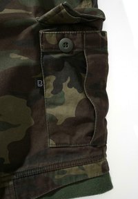 Karinis audinys žalios, rudos ir tamsiai smėlio spalvos; turi didelį kišenę su mygtuko uždarymu ir sulankstytą kraštą, lygios tekstūros.