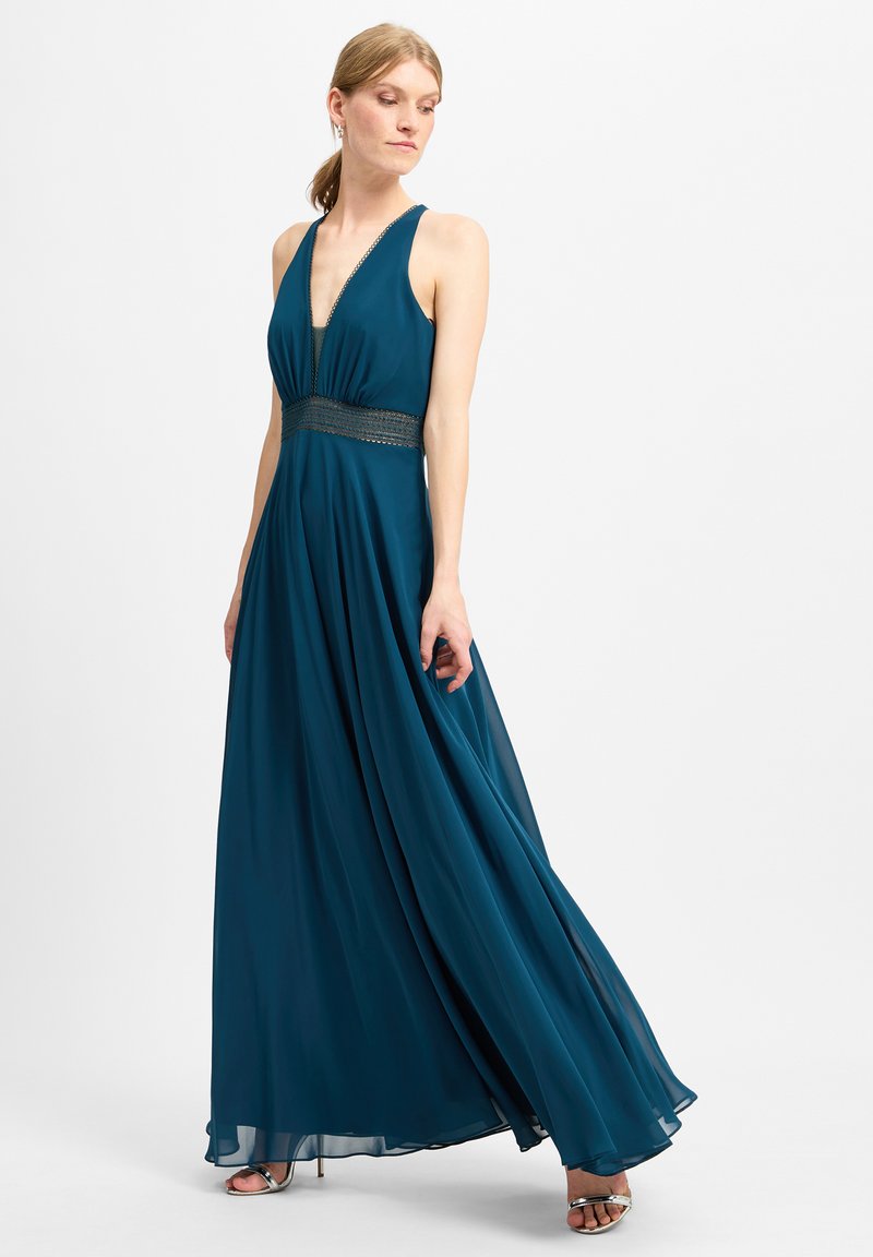 Tannengrünes Maxikleid mit tiefem Ausschnitt, geraffter Taille und metallischen Details. Fließender Stoff erzeugt eine sanfte Silhouette.