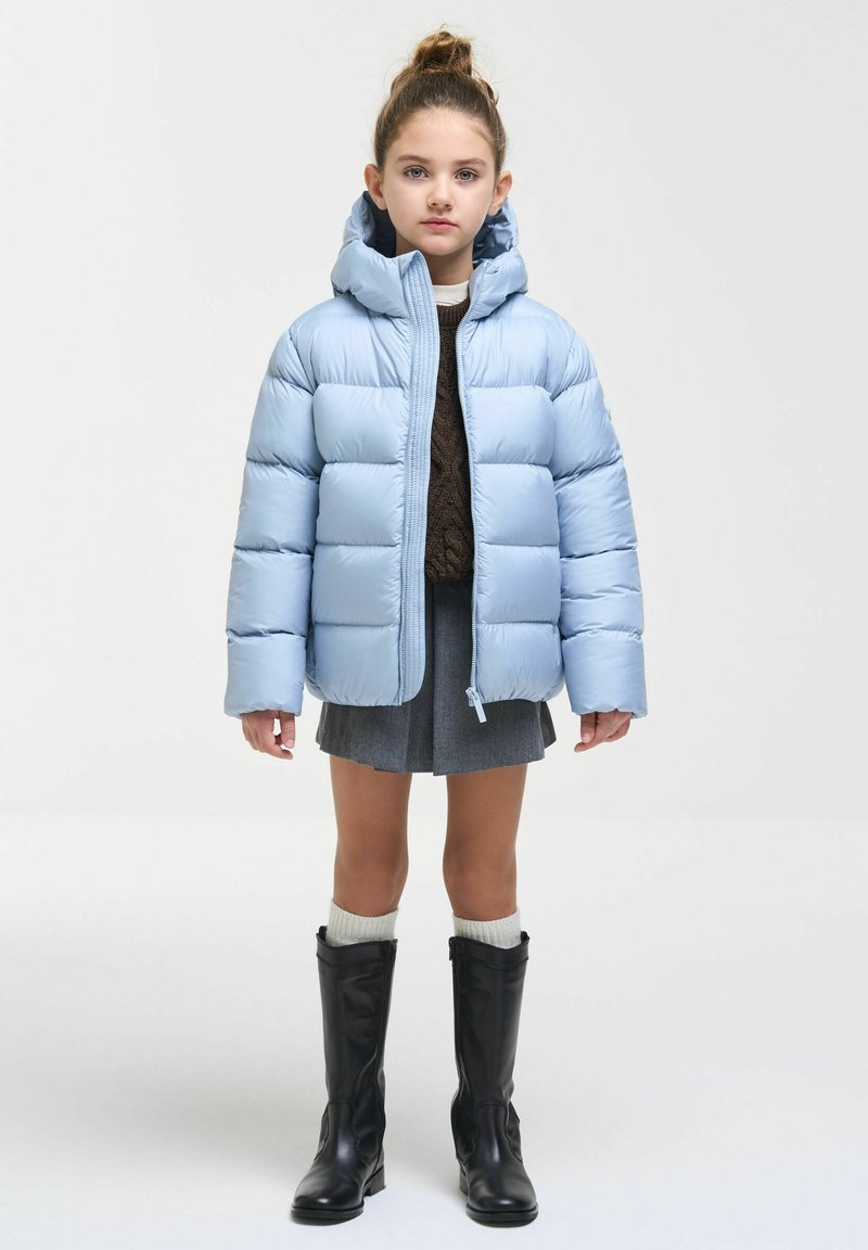 Jeune fille avec les cheveux attachés en chignon, portant une doudoune bleu clair, une jupe grise, un pull marron, des chaussettes blanches et des bottes noires montant jusqu'aux genoux, debout sur fond blanc.