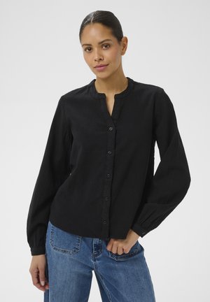 Tumejuustega patsiga naine, kes kannab musta pika varrukaga nööbitavat pluusi ja siniseid teksaseid, seisab valgel taustal.