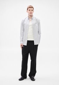 Πουκάμισο με ρίγες σε ανοιχτό γκρι και λευκό, φορεμένο πάνω από κρεμ αμάνικο τοπ, συνοδευόμενο από άνετο μαύρο παντελόνι και μαύρα παπούτσια.