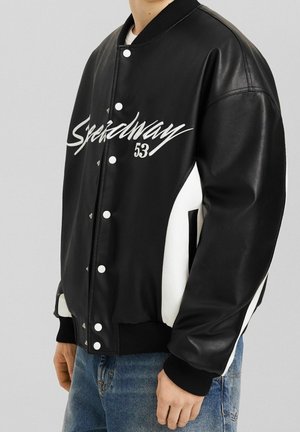 Person wearing a black and white varsity jacket with "Speedway 53" text, paired with blue jeans, standing against a gray background.