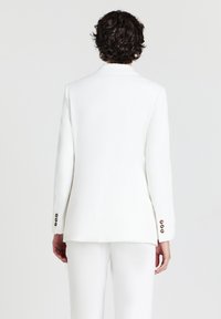 Blazer bianco su misura con una vestibilità strutturata, dotato di colletto a punta e polsini con bottoni. Texture liscia, design minimalista senza decorazioni.