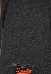 Tela de camiseta de color gris oscuro con una textura suave; cuenta con una pequeña etiqueta roja "SuperDry" cosida en el dobladillo inferior.