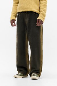 Persoană îmbrăcată cu blugi negri largi, adidași verzi și albi și un pulover tricotat galben, stând în fața unui fundal alb.