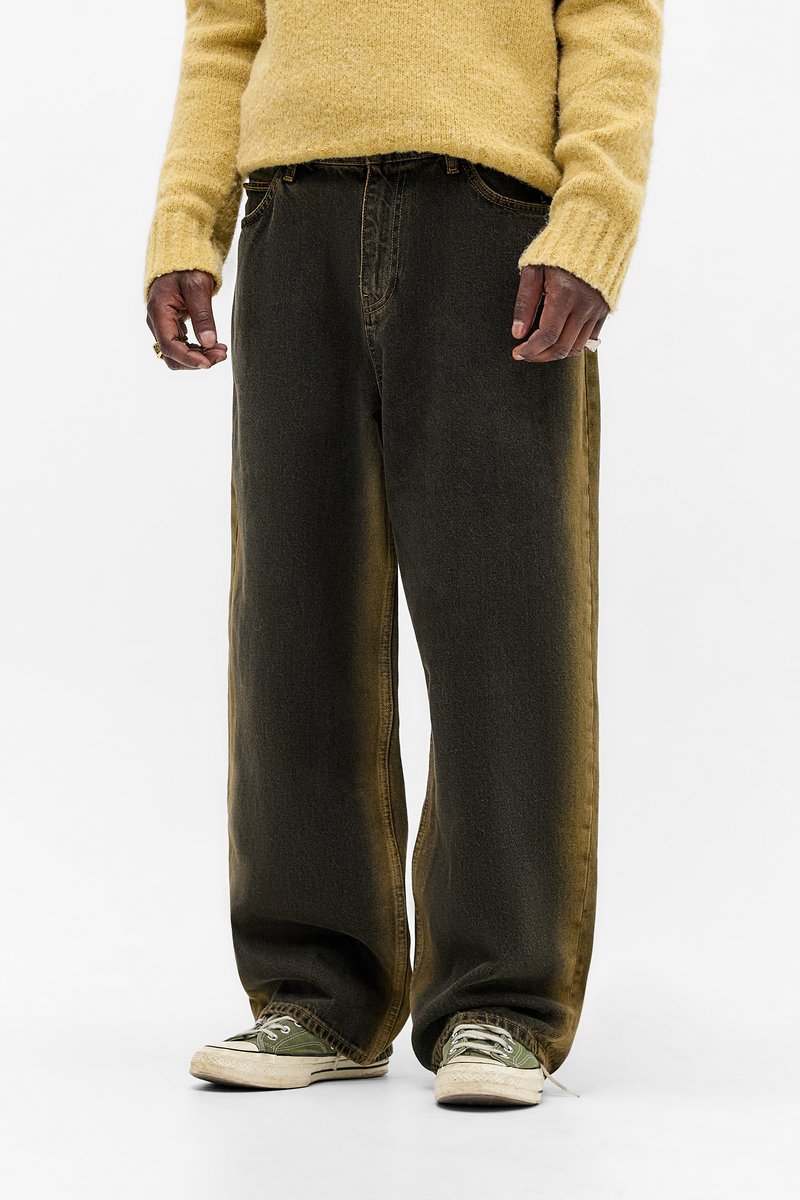 Persoană îmbrăcată cu blugi negri largi, adidași verzi și albi și un pulover tricotat galben, stând în fața unui fundal alb.