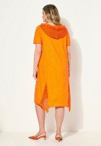 Orange Kleid mit kurzen Ärmeln, Kapuze mit Spitzenbesatz und seitlichen Schlitzen. Getragen mit orangenen Slingback-Heels, durchgehend aus strukturiertem Stoff.