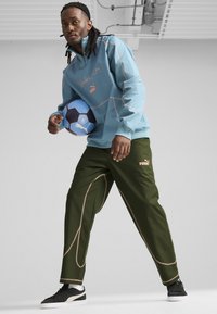 Puma ceturtdaļas rāvējslēdzēja jaka gaiši zilā krāsā ar kontrastējošām šuvēm, kopā ar olīvzaļām biksēm ar dekoratīvo šuvi. Melni sporta apavi un zila futbola bumba.