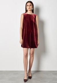 Sukienka bez rękawów w głębokim burgundowym aksamicie, z okrągłym dekoltem i subtelnymi bocznymi kieszeniami, w połączeniu z czółenkami o spiczastym nosku.