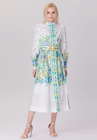 Robe maxi florale en tissu blanc transparent, ornée de motifs bleus et jaunes, avec un col haut, de longues manches bouffantes et une ceinture contrastante à boucle ronde.