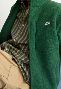 Vihreä fleece-takki vetoketjulla, jossa valkoinen Nike-logo. Alla vihreä-valkoraidallinen nappipaita ja vaaleat khakinväriset housut.