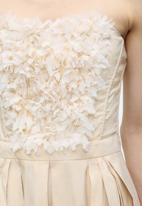 Κρεμ φόρεμα χωρίς τιράντες με ανάγλυφες υφασμάτινες εφαρμογές λουλουδιών και μικρές στρας στο μπούστο, με πλισέ φούστα κάτω από τη μέση.