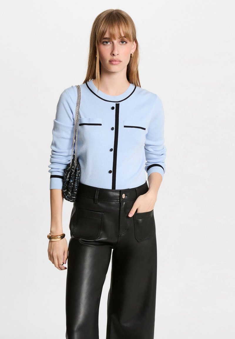 Cardigan lavorato a maglia blu chiaro con accenti neri, caratterizzato da un collo rotondo, bottoni sul davanti e due tasche sul petto. Abbinato a pantaloni in pelle nera.