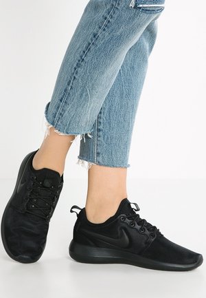 Stopy w czarnych trampkach Nike z sznurowadłami, w połączeniu z krótkimi, postrzępionymi niebieskimi dżinsami, ustawione na jednolitym białym tle.