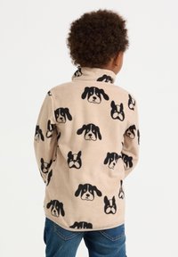 Bambino che indossa una giacca in pile beige con motivi a faccia di cane nera, in piedi con le spalle alla telecamera contro uno sfondo bianco semplice.