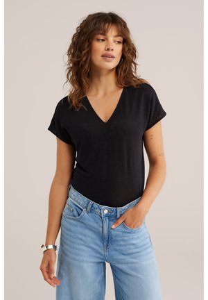 Kobieta o kręconych brązowych włosach, ubrana w czarną koszulkę z dekoltem w serek i jasnoniebieskie dżinsy, stojąca z jedną ręką w kieszeni na neutralnym tle.