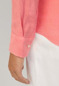 Manica di camicia in lino rosa con dettaglio di bottone, caratterizzata da un design a polsino e una leggera trama, abbinata a tessuto bianco sottostante.