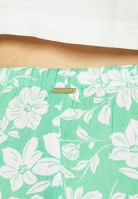 Tissu à motif floral vert avec des fleurs blanches et une taille froncée. Etiquette en métal avec "Billabong" fixée à la taille.