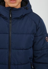 Marineblå polyesterjakke med høj krave, kviltet mønster, dekorativ lynlås og logo-patch på skulderen. Har en glat, blød tekstur.