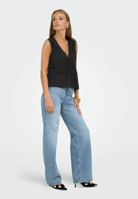 Zwarte mouwloze top met een V-hals, voorzien van bloemenborduursels; gecombineerd met lichtblauwe wijde jeans en zwarte puntige hakken met stippen.