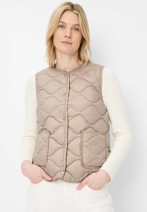 Blonde Frau, die eine beige gesteppte Weste mit Fronttaschen über einem weißen Langarmshirt trägt, steht vor einem weißen Hintergrund.