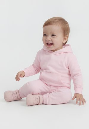 Kisgyermek ülve és mosolyogva, világos rózsaszín kapucnis pulóvert, hozzáillő melegítőnadrágot és rózsaszín tornacipőt viselve, fehér háttér előtt.