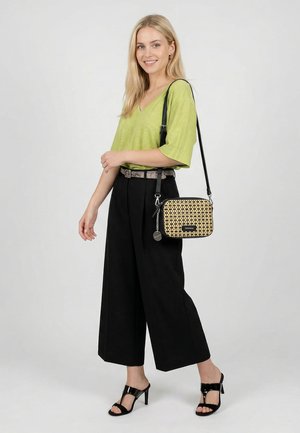 Ung kvinde smiler, iført lys grøn top, sorte vidde bukser, sorte hælede sandaler og bærer en lille mønstret crossbody taske.