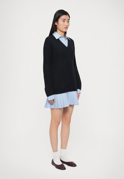 Fekete kötött pulóver V-nyakkivágással, világoskék csíkos galléros ruhával párosítva. Fehér zoknival és bordó balerinacipővel viselve.