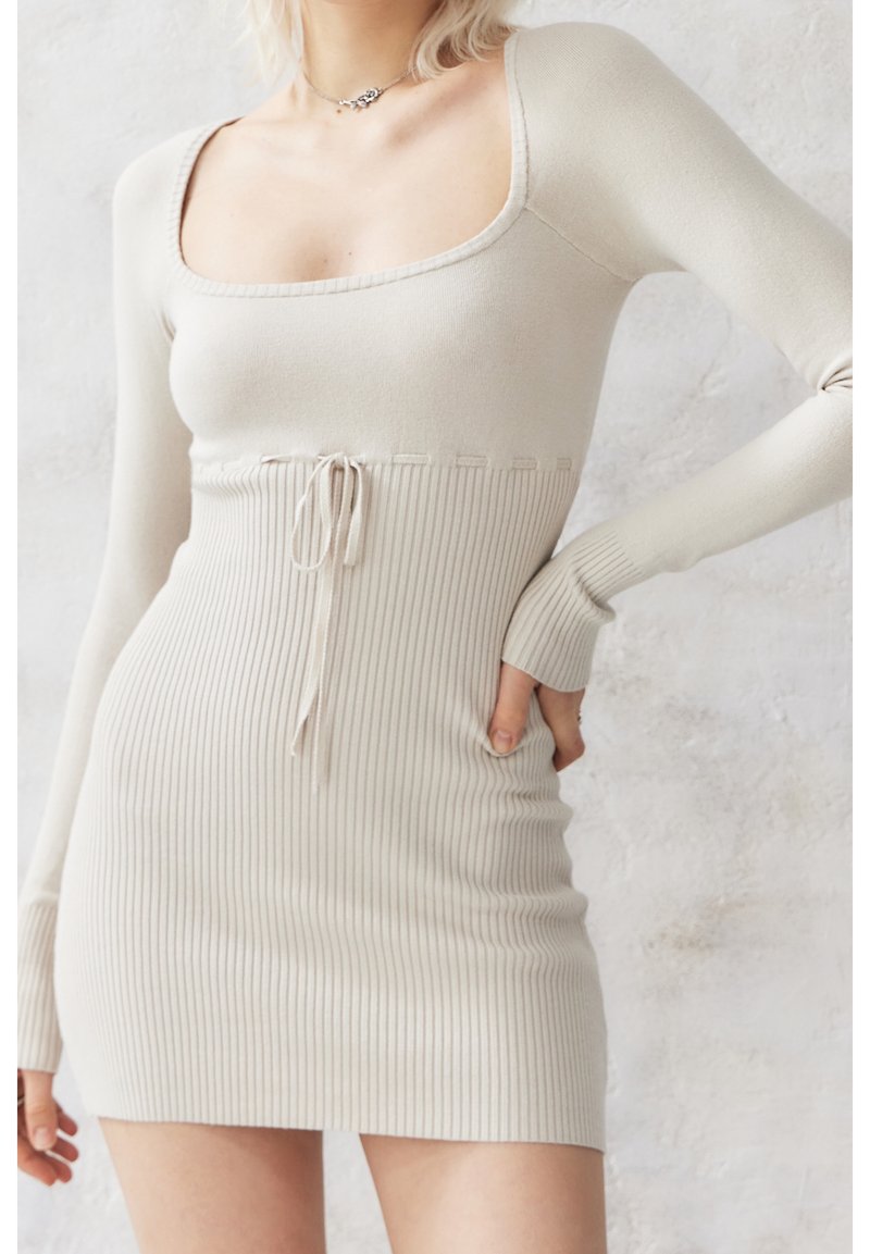 Femme portant une robe en maille côtelée beige à manches longues, col carré et détail noué à la taille, debout devant un fond clair texturé.