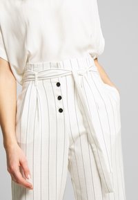 Vita, högmidjade byxor med svarta vertikala ränder, med bälte i midjan, fyra knappar och sidfickor. Slät tygkvalitet.