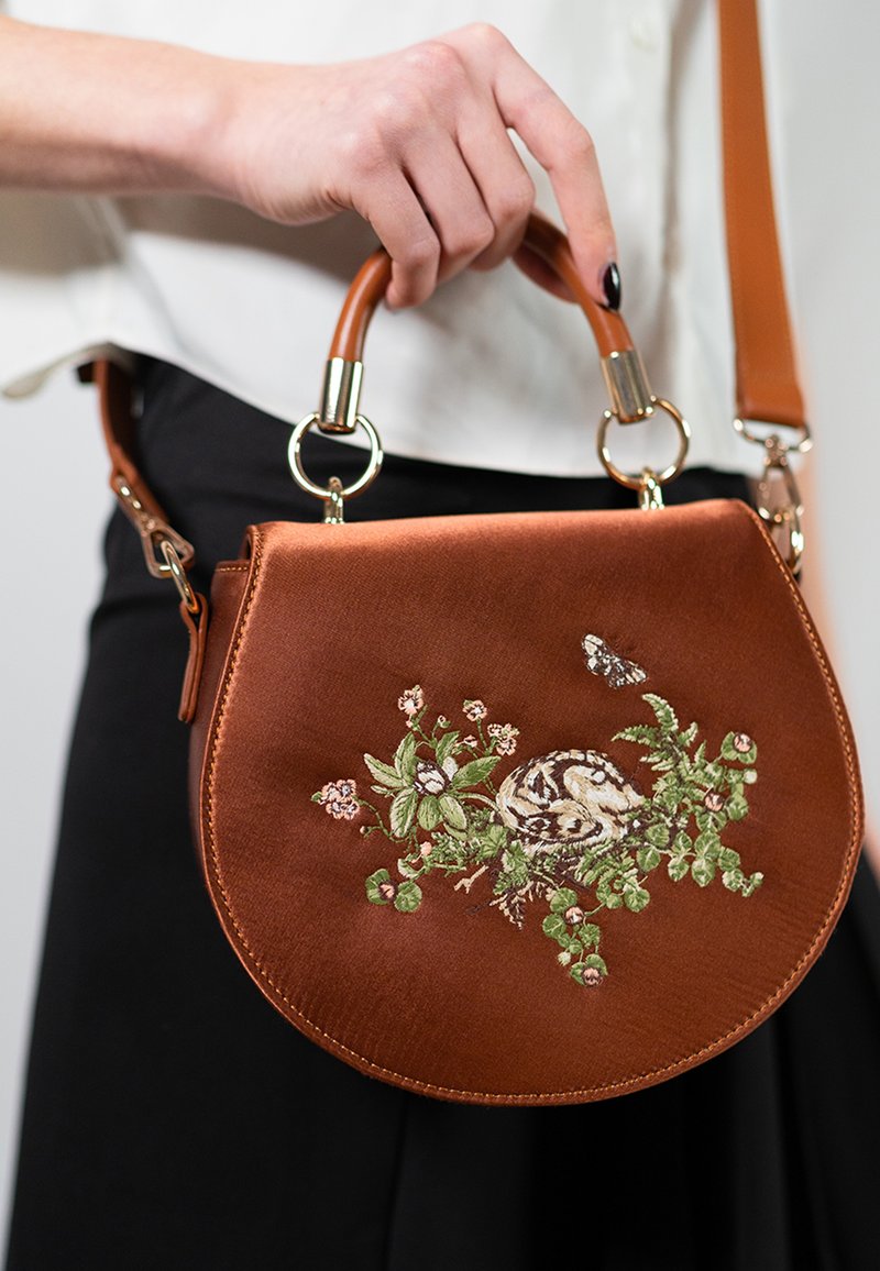 Brown circular handbag made of textured material, featuring floral embroidery with a central design, metal accents, and a detachable shoulder strap.