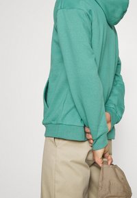 Felpa turchese con tasca frontale, polsini a costine e cappuccio con coulisse, abbinata a pantaloni beige, con una mano che tiene un cappello beige.