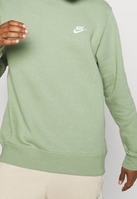 Grön bomullströja med rund hals, ribbade ärmslut och nederkant. Har en vit Nike-logotyp på vänster bröst. Mjukt, texturerat tyg.