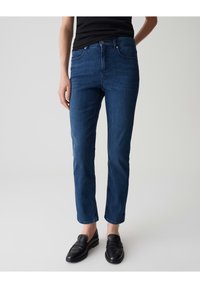 Jeans en denim bleu foncé avec une coupe slim, représentant un design classique à cinq poches et une fermeture à bouton. Associés à des mocassins noirs.