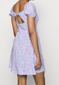 Robe florale en tissu bleu clair avec des fleurs roses et blanches. Présente des manches bouffantes et un dos découpé avec un détail à nouer. Silhouette évasée.