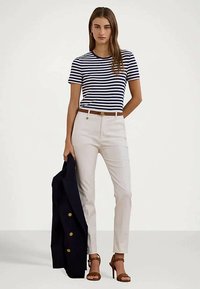 Γυναίκα ντυμένη με μπλε και λευκή ριγέ μπλούζα, κρέμα παντελόνι με καφέ ζώνη, καφέ τακούνια, κρατώντας μπλε σακάκι με χρυσά κουμπιά.