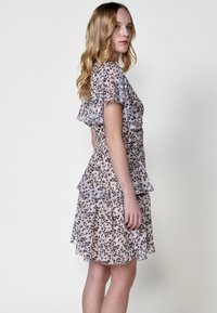 Blumendruckkleid mit geschichteten Rüschen, kurzen Ärmeln und tailliertem Oberteil mit gerüschtem Bund. Die Farben sind sanftes Beige mit schwarzen Akzenten.