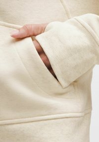 Kremasti dukserica s teksturiranim materijalom, rebrastim manžetama i prednjim džepom s glatkim rubom. Krupni kadar ruke koja drži manžetu.
