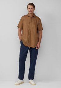 Korte mouwen bruin linnen shirt gecombineerd met marineblauwe broeken en witte sneakers. Het shirt heeft knopen en een relaxed fit. Glad oppervlak gedurende het hele shirt.