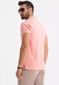 Rosa pikétröja med korta ärmar, krage, sidoflikar och en jämn textur, ihop med beiga byxor och mörka solglasögon.