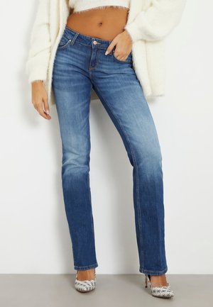 Kobieta ubrana w niebieskie dżinsy o prostych nogawkach z podwyższonym stanem, biały puszysty krótki sweter oraz białe wzorzyste szpilki, stoi z jedną ręką w kieszeni.