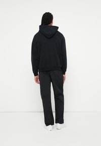 Črna kapucna majica ohlapnega kroja, v kombinaciji s črnimi trenirkami z belimi stranskimi črtami, obuta z belimi športnimi čevlji.