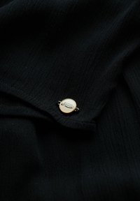 Tela negra con textura, con un pin redondo en tono dorado que lleva el logo de "Superdry" en un lado.