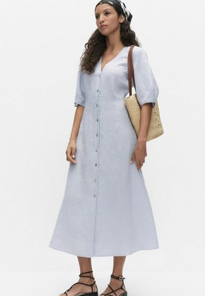 Kvinde iført en lyseblå midi-kjole med knapper foran, sorte sandaler med stropper, et sort-hvidt tørklæde om hovedet og bærende en flettet stråtaske over den ene skulder.