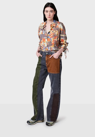 Donna che indossa una camicia a maniche lunghe con stampa astratta colorata e jeans a gamba larga in patchwork con sneakers, in piedi con le mani nelle tasche.
