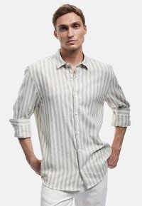 Camisa ligera de lino en color beige con rayas blancas verticales. Cuenta con un frente abotonado, mangas enrolladas y un dobladillo curvado.