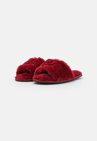 Une paire de chaussons ouverts en rouge duveteux avec des semelles noires sur un fond blanc, légèrement inclinés vers la gauche.