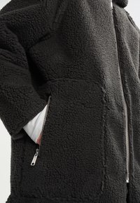 Musta fleecetakki, jossa on teksturoitu pinta, edessä vetoketju ja sivutaskut. Kangas näyttää pehmeältä ja paksulta.