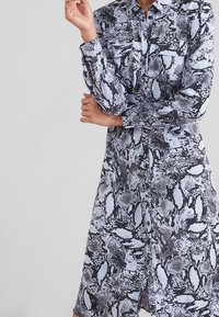 Langärmliges, kragenloses Hemdkleid in Hellblau mit einem Schwarz-Weiß-Schlangenmustermuster. Verfügt über eine Knopfverschluss und zwei vordere Taschen.