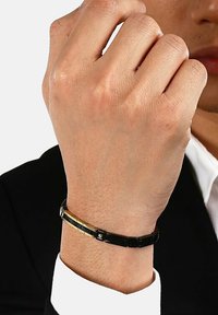Svart metallarmband med guldaccenter, med en elegant design och slät yta, bärs på ett handled mot en mörk kostym.